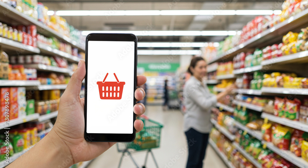 Fototapeta premium Person holding smartphone with shopping basket icon while shopping in a grocery store.