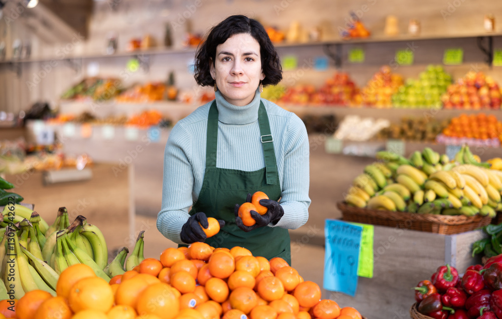 Shop seller puts goods on display case. Woman creates pyramid of tangerine, presents fresh goods. Vegetable shop near house. Spacious store with long rows of display cases, customers waiting Shop seller puts goods on display case. Woman creates pyramid of tangerine, presents fresh goods. Vegetable shop near house. Spacious store with long rows of display cases, customers waiting