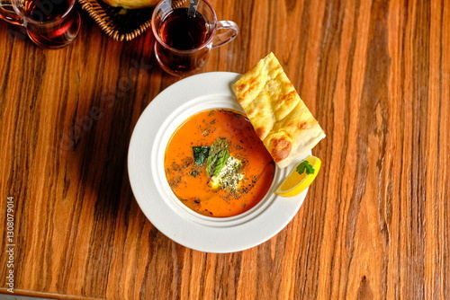 Turkish Tarhana soup with meatballs in a white bowl, garnished with herbs, lemon, bread, and tea on a wooden table.
