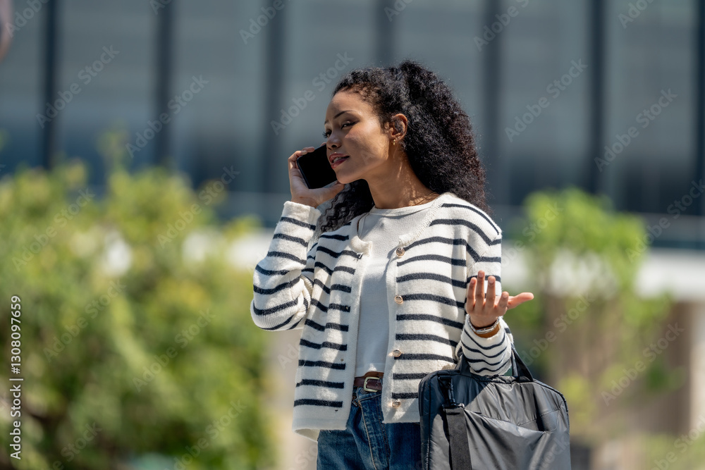 woman talking on smartphone while holding bag, wearing striped sweater and jeans, outdoors in city setting. She appears engaged and expressive woman talking on smartphone while holding bag, wearing striped sweater and jeans, outdoors in city setting. She appears engaged and expressive
