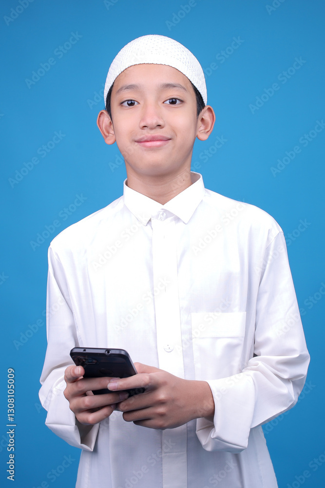 Young Boy Wearing Muslim Clothes Smiling While Using Smartphone Isolated On Blue Background Young Boy Wearing Muslim Clothes Smiling While Using Smartphone Isolated On Blue Background