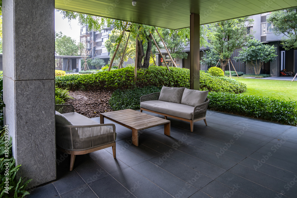 Obraz premium A modern outdoor patio of a residential apartment complex features stylish seating with chairs and a coffee table, against manicured shrubs.The space covered by a sleek roof, overlooking a courtyard.