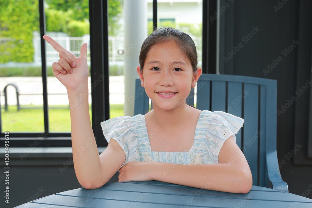 Smiling Asian girl child show index finger up while sitting on chair indoor.