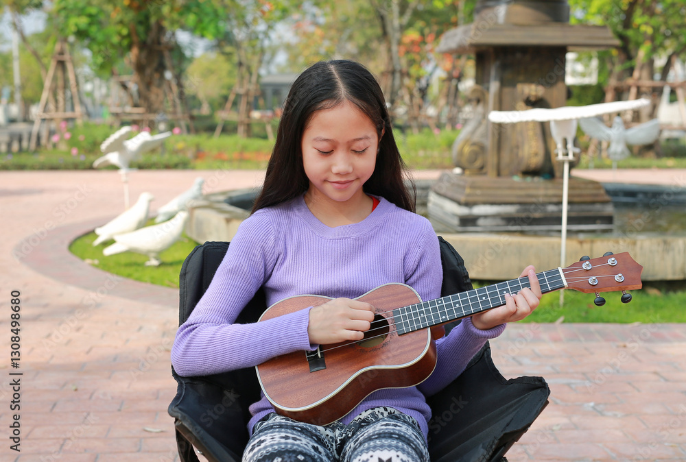 Portrait of Asian pretty girl playing ukulele while sitting on canvas chair in the garden.