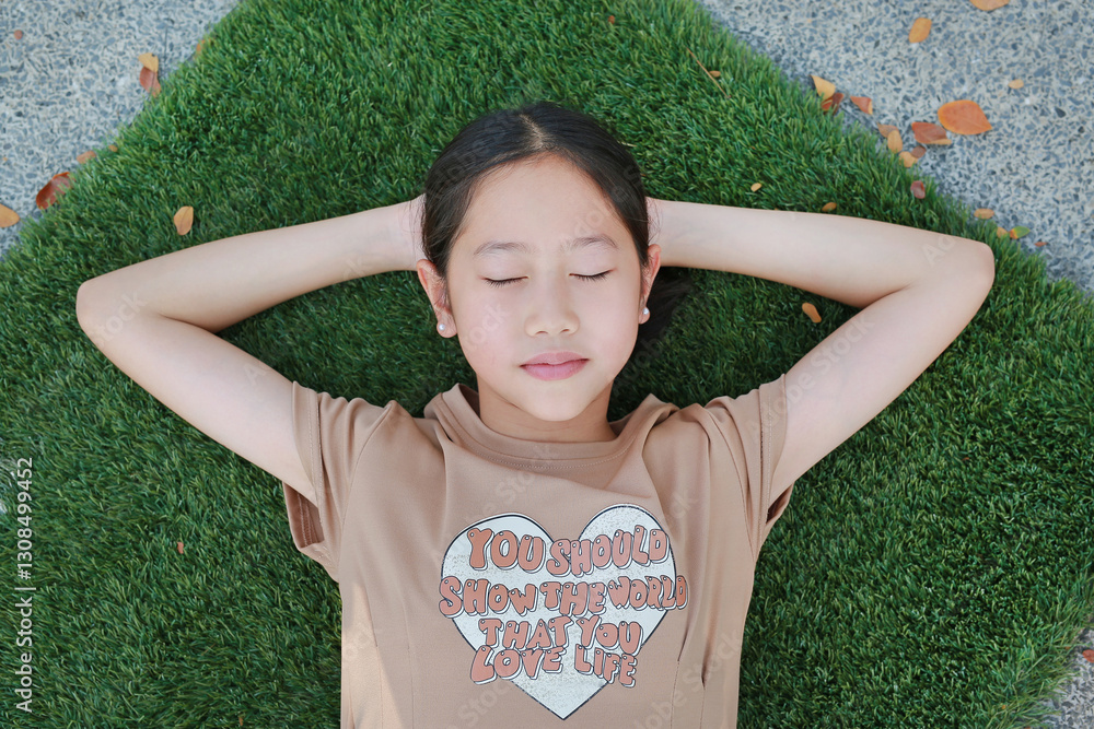 Close-up top view of Asian girl child lying on green grass outdoors in summer park. Kid closed eyes resting in garden.
