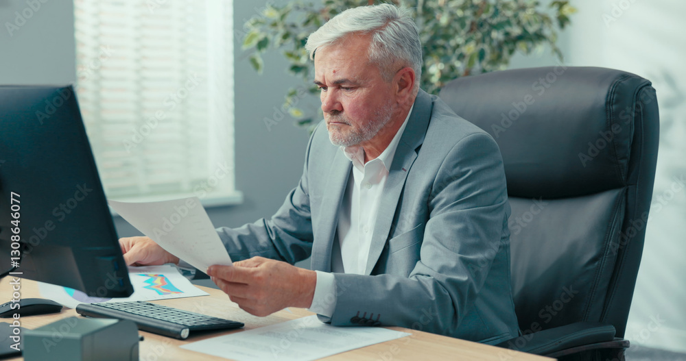 Fototapeta premium Mature, gray-haired, elegant businessman sits in office at the computer, completes company documents, checks data in the system, sorts invoices, performs managerial duties
