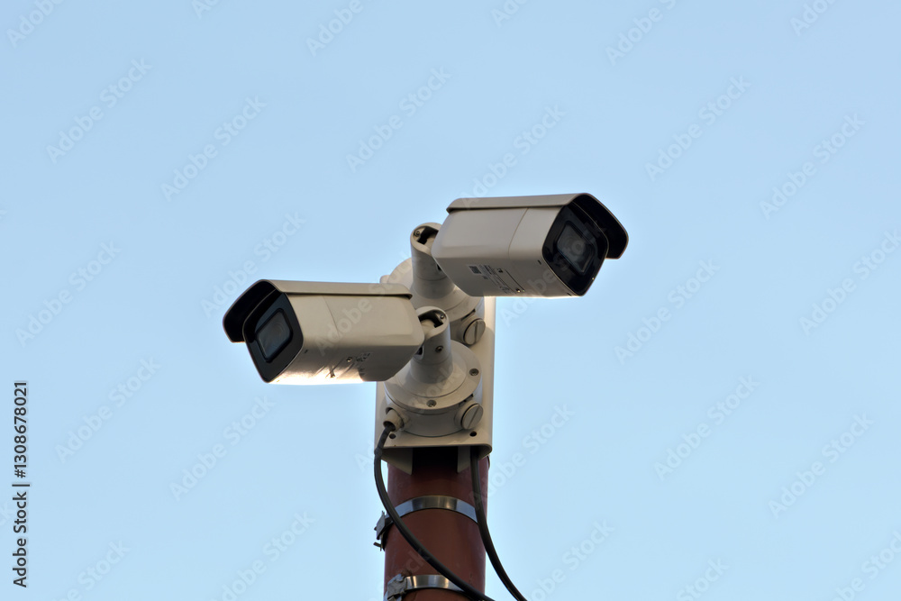 Two surveillance cameras mounted on a pole against a clear blue sky, representing security, monitoring, and urban surveillance systems. Two surveillance cameras mounted on a pole against a clear blue sky, representing security, monitoring, and urban surveillance systems.