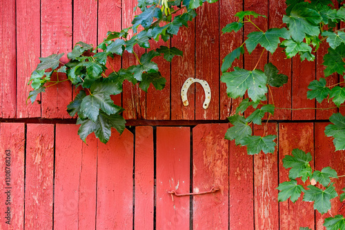 A rustic red barn door features a horseshoe and is surrounded by lush green vines on a serene countryside backdrop.