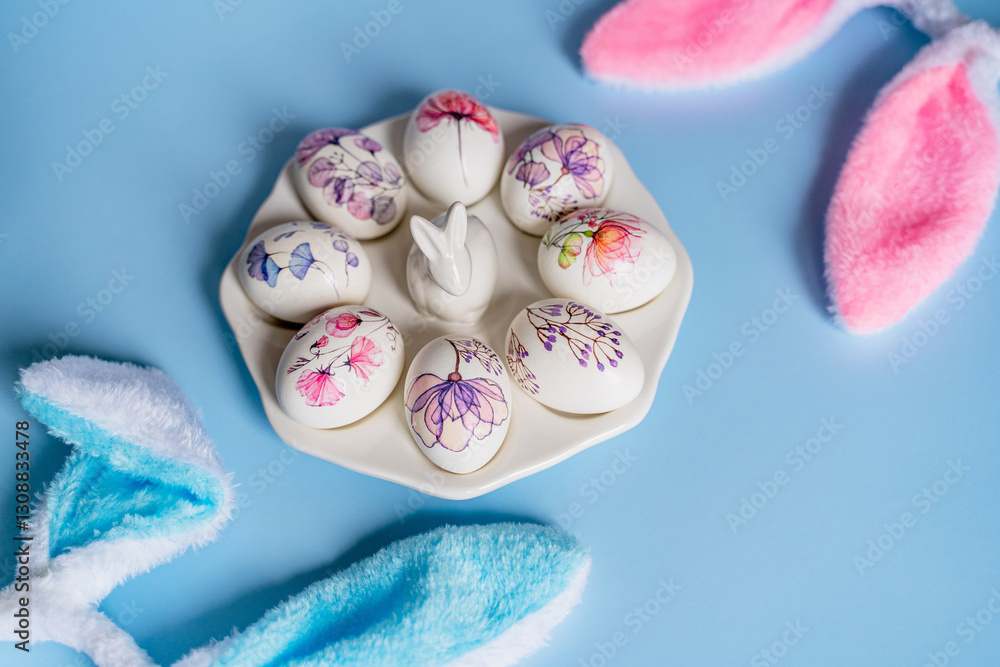 Easter beautiful decorated orthodox religious chicken eggs with a picture of flowers lie on a white porcelain plate with a porcelain rabbit on a blue background and with fur rabbit ears and space for Easter beautiful decorated orthodox religious chicken eggs with a picture of flowers lie on a white porcelain plate with a porcelain rabbit on a blue background and with fur rabbit ears and space for