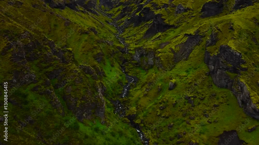 Aerial drone flying over a narrow river flowing through mossy steep mountains and cliffs during a cloudy day in Iceland. The cliffs are steep, with rough edges and high with greenery and wet moss.