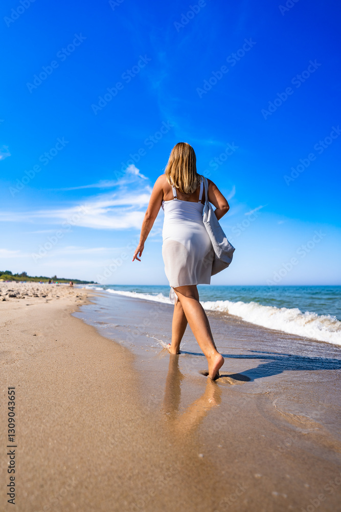 Summer vacation by sea. Beautiful elegant middle aged woman tourist with beach bag walking along sea on sandy beach on sunny day. Back view Summer vacation by sea. Beautiful elegant middle aged woman tourist with beach bag walking along sea on sandy beach on sunny day. Back view