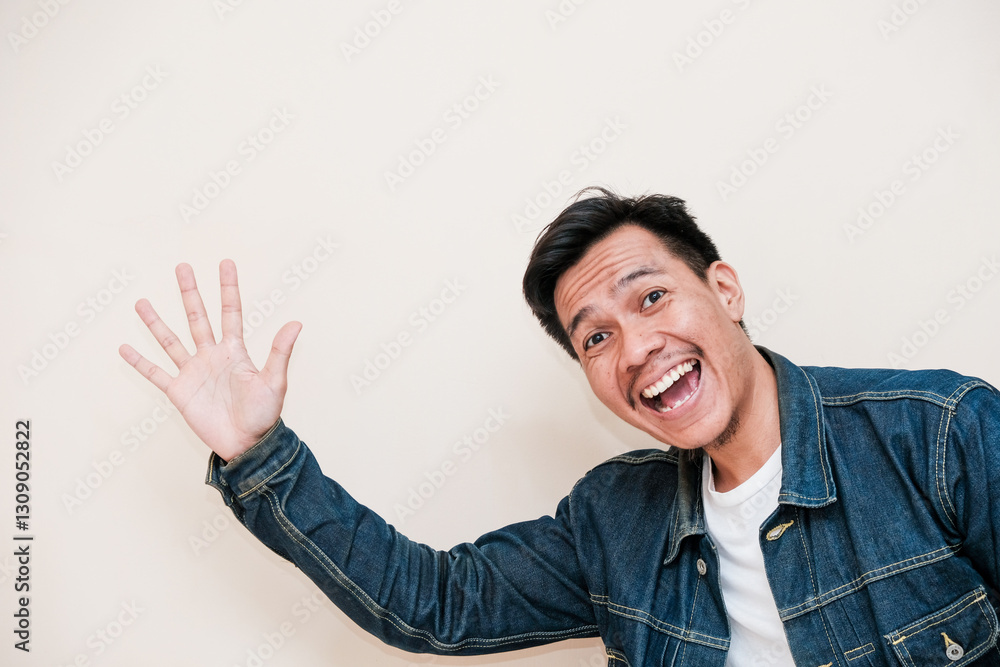 A Southeast Asian man wearing a denim jacket smiling pop out for greeting and say hi against a white background. This image depicts greeting A Southeast Asian man wearing a denim jacket smiling pop out for greeting and say hi against a white background. This image depicts greeting