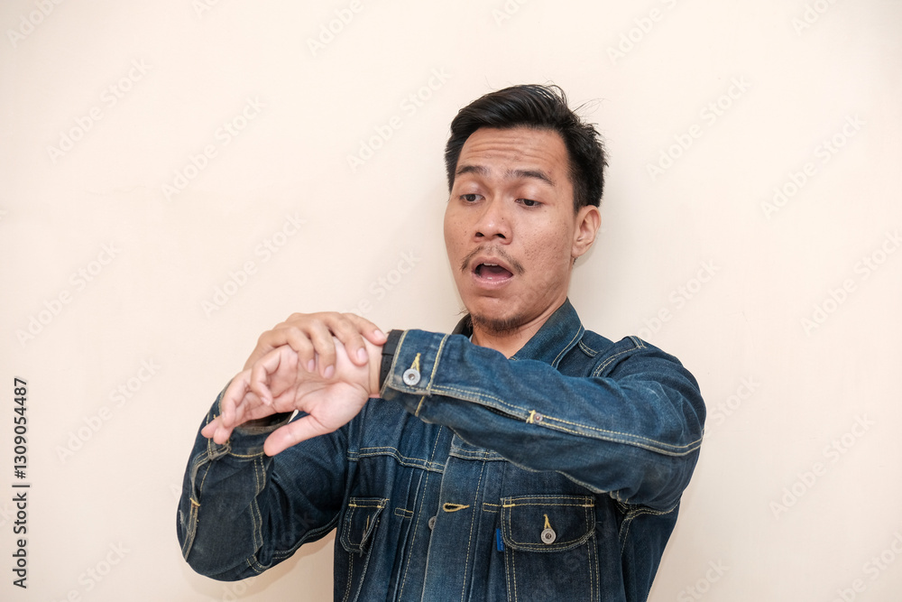 Obraz premium A Southeast Asian man wearing a denim jacket shocked expression to see watch on hand against a white background. This image is perfect for content related to time management