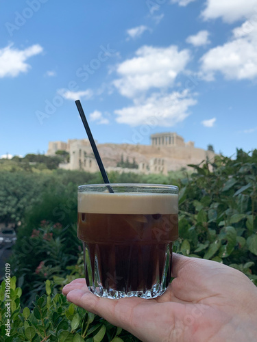 Hand holding a glass of freddo espresso with a black straw against the backdrop of the Acropolis in Athens.