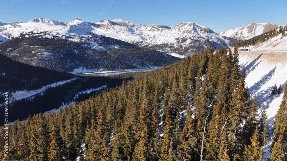 Aerial view of snow-covered mountain peaks in Colorado, with dense evergreen forests, rugged alpine terrain, and a clear blue sky highlighting the winter landscape