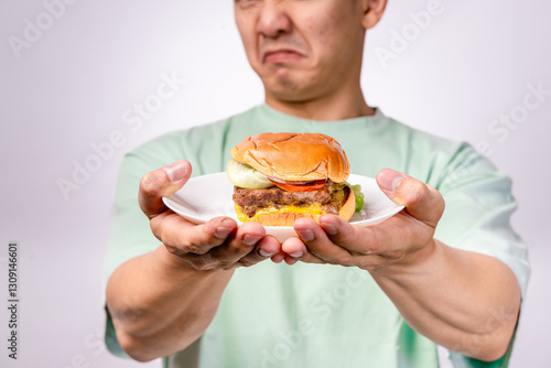 An Asian man in a mint green shirt holds a plate with a cheeseburger while making a disgusted expression. The background is white