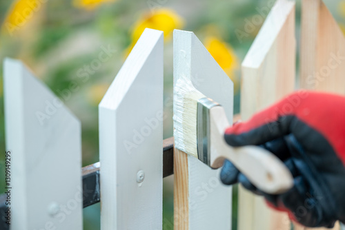 Close up of man hand in protective glove painting fence in white color. Painting new wooden fence with a brush in white color