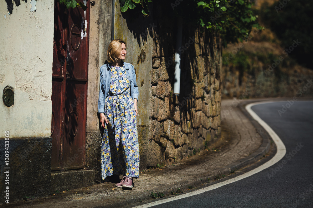 A woman in a light dress stands near a road in a picturesque Portuguese village, exuding charm and simplicity.
