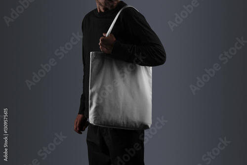 A tote bag mockup, carried by a man dressed in black attire, including a long-sleeved shirt, and pants. Isolated blank mockup surface to place your design. 