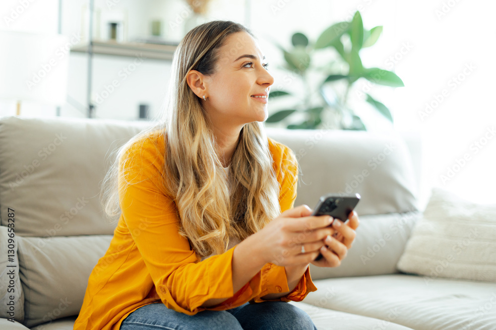 Beautiful woman is sitting on a couch, holding a smartphone, looks happy while using her mobile phone in a cozy home environment, chatting or browsing social media, looking away and smiling Beautiful woman is sitting on a couch, holding a smartphone, looks happy while using her mobile phone in a cozy home environment, chatting or browsing social media, looking away and smiling