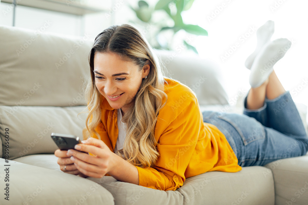 Cheerful caucasian pretty woman is lying on a sofa at living room, holding a smartphone, messaging online, reading news, making a payment or shopping online, browsing social media, smile Cheerful caucasian pretty woman is lying on a sofa at living room, holding a smartphone, messaging online, reading news, making a payment or shopping online, browsing social media, smile
