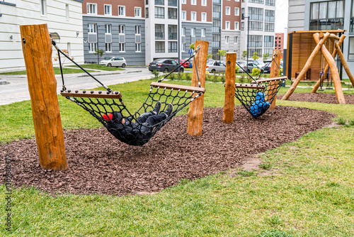 Kids lying on black net hammocks in a modern residential playground with wooden structures, surrounded by green grass and apartment buildings