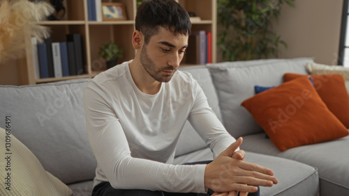 Fotografie Young man with contemplative expression sits in a cozy living room, surrounded by plush pillows and a book-filled shelf, embodying a peaceful and introspective atmosphere
