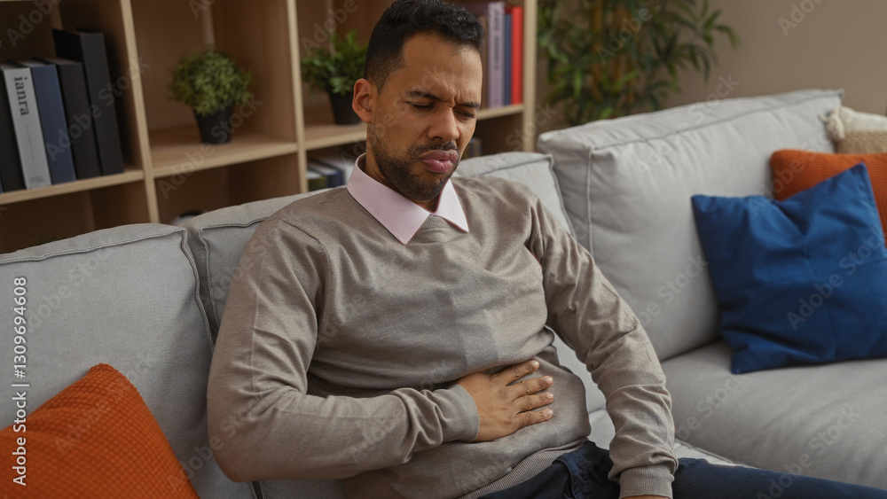 Man indoors holding stomach in discomfort on a sofa in a cozy living room with modern furnishings implying a casual home setting. Man indoors holding stomach in discomfort on a sofa in a cozy living room with modern furnishings implying a casual home setting.