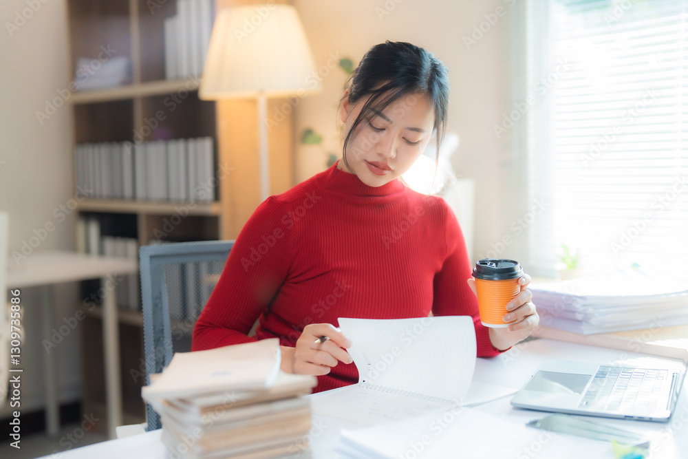 Young Asian businesswoman dressed in a vibrant red turtleneck sweater, reviewing important documents and sipping coffee while working at her desk in a sleek, modern office environment