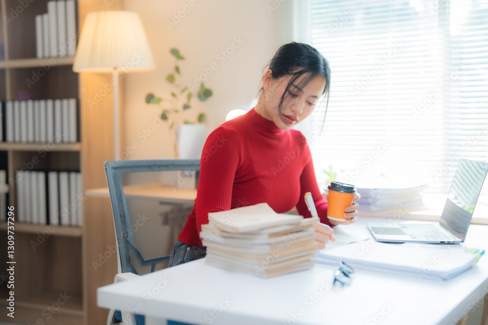 Young Asian businesswoman sitting at her desk working from her home office, taking notes from a pile of books and holding a cup of coffee, with laptop and paperwork also on the desk Young Asian businesswoman sitting at her desk working from her home office, taking notes from a pile of books and holding a cup of coffee, with laptop and paperwork also on the desk