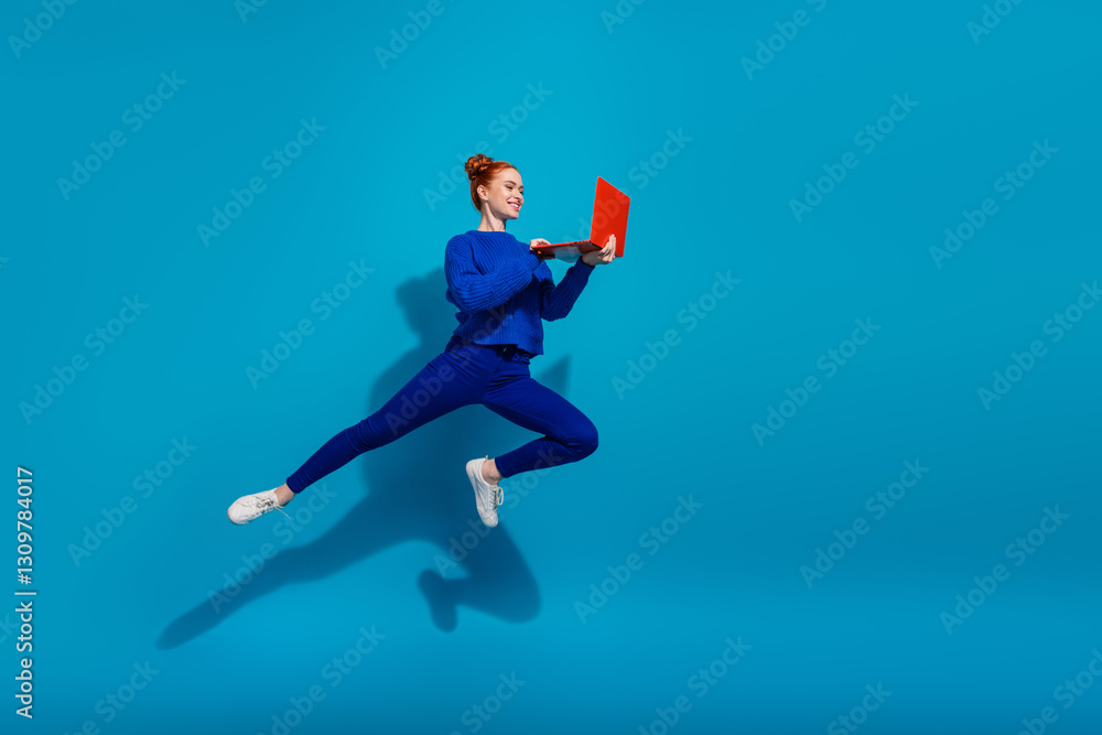 Fototapeta premium Young woman with red hair jumping energetically on blue background holding a laptop, radiating energy and joy