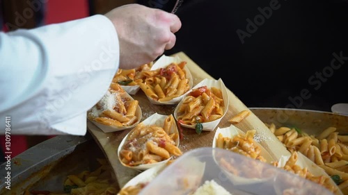 Professional chef in a white uniform sprinkles grated cheese over servings of penne pasta in disposable plates. Close-up of hands adding final touches at a catering event or professional kitchen.