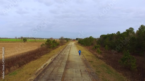 Aerial view of a man wearing a blue track suit running alone down an old unmaintained rural roadway beside a forest and farmland on a cloudy day