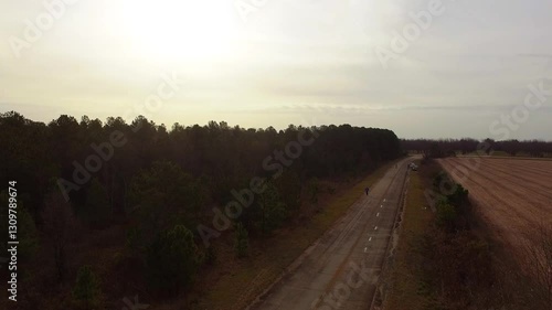 Aerial view of a man wearing a blue track suit jogging along an old unmaintained rural roadway beside a forest and farmland on a cloudy day