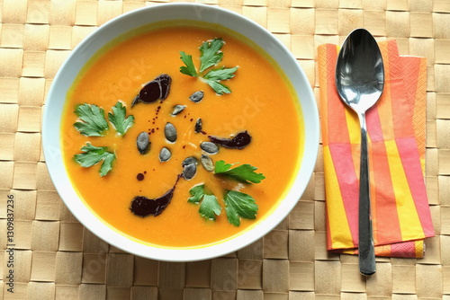 Wallpaper Mural Ein Teller Kürbissuppe mit Petersilienblättern, Kürbiskernöl und Kürbiskernen drauf auf einem hellorangefarbenen Untersetzer und neben einer Serviette und einem Esslöffel aus Edelstahl. Torontodigital.ca