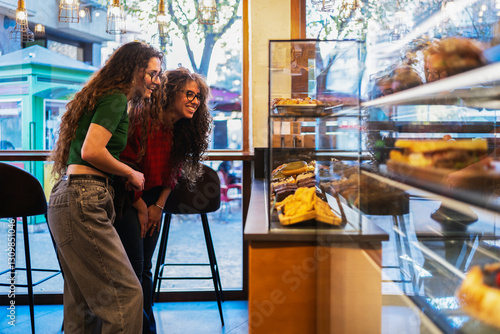 Two young women having fun selecting delicious pastries from a display case in a bright, modern cafe