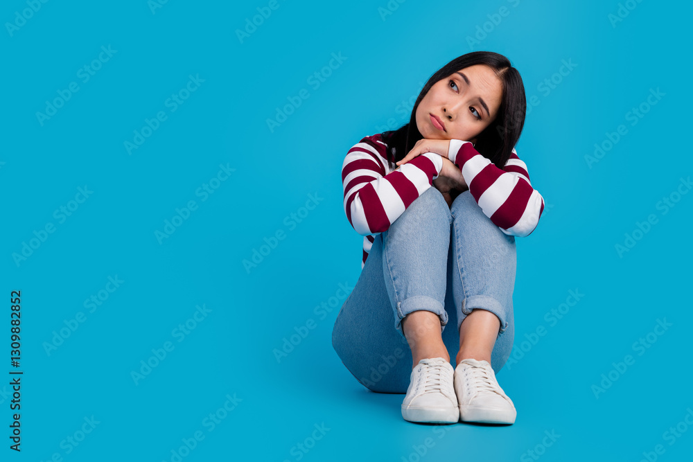 Young thoughtful woman sitting against blue background wearing casual striped shirt and jeans Young thoughtful woman sitting against blue background wearing casual striped shirt and jeans