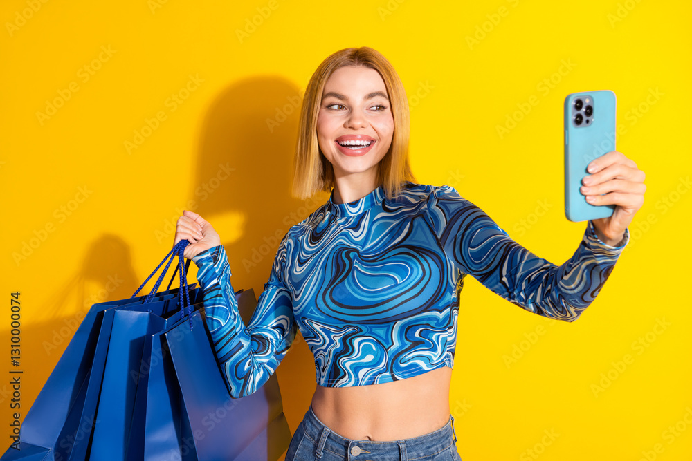 Φωτογραφία Beautiful young woman with blond hair enjoying shopping and taking a selfie, holding blue bags, against a yellow background