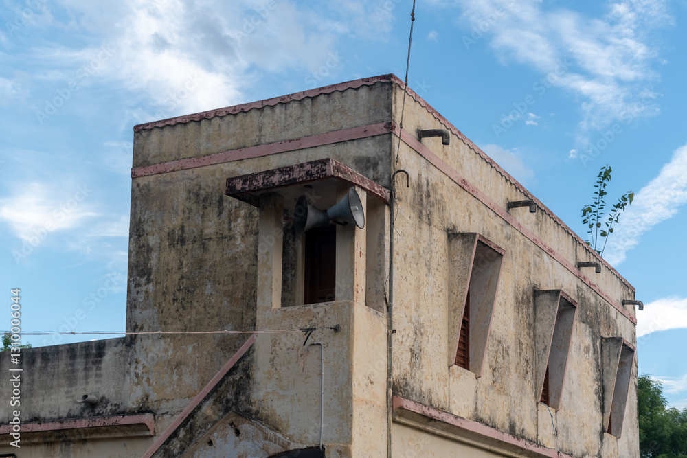 Fototapeta premium A weathered building stands against a bright blue sky, featuring speakers mounted on its wall.