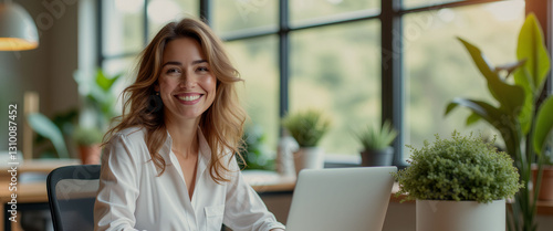 A woman is sitting at a desk with a laptop and a potted plant in front of her. She is smiling and she is happy