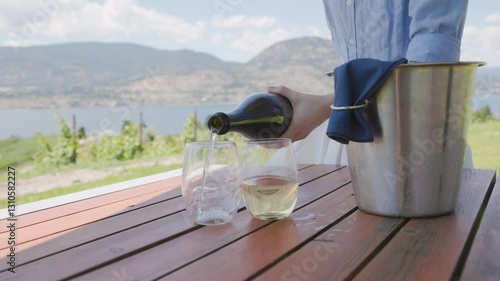Woman's hand pouring bottle of sparkling wine champagne in glasses on patio overlooking beautiful lake country vista, Okanagan, wine, vineyard. 4K 24FPS