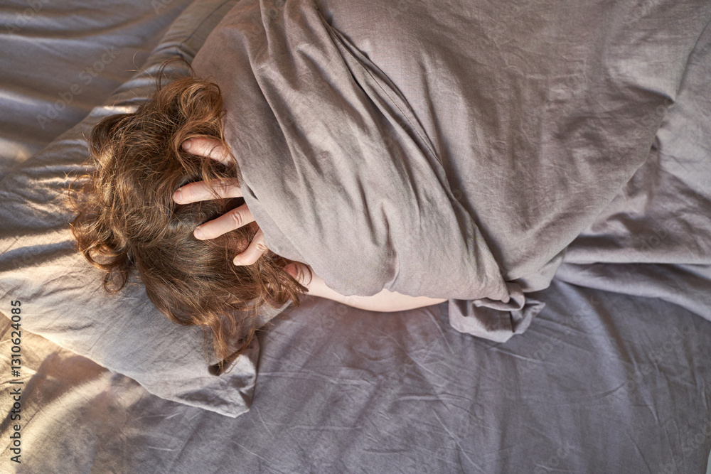 Obraz premium Female caucasian adult wrapped in grey blanket lying in bed with hands on head in relaxed morning mood.