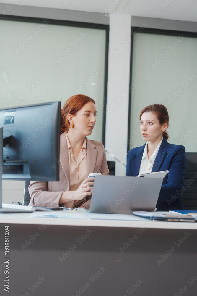 Two Caucasian women work in an office, happily using laptops and computers at a desk, discussing financial projects, analyzing financial statements,collaborating in a meeting to ensure effective
