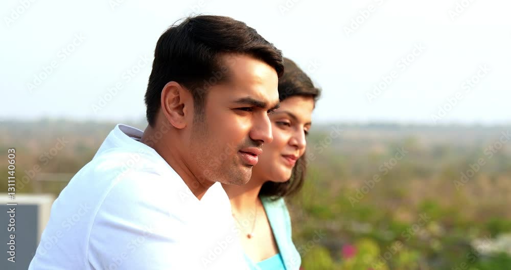 Indian Asian young beautiful happy couple standing on balcony at railing, chatting, laughing, pointing at something far away, enjoying a relaxed morning together drinking coffee outdoors in fresh air Indian Asian young beautiful happy couple standing on balcony at railing, chatting, laughing, pointing at something far away, enjoying a relaxed morning together drinking coffee outdoors in fresh air