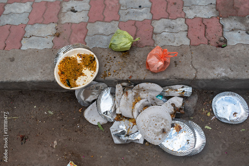 Food waste and discarded plates clutter a busy street in India, highlighting the aftermath of a local festival. Litter includes leftover curry and packaging, creating a scene of neglect.