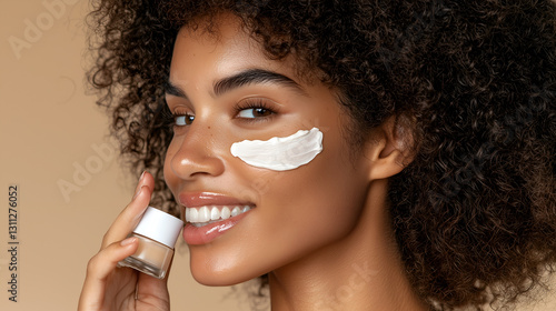 A smiling woman applies a skincare mask to her face, showcasing healthy skin and curly hair against a warm, neutral background.