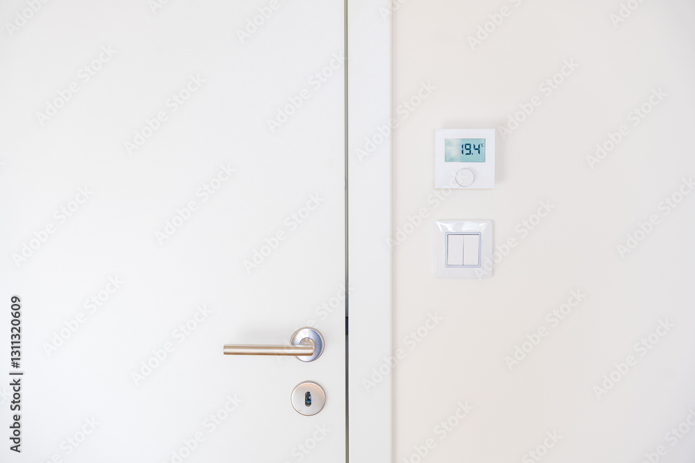 Minimalistic interior with a modern white door featuring a stainless steel handle and lock. A digital thermostat displays 19.4 above a light switch. Captured from a straight-on angle. No people. Minimalistic interior with a modern white door featuring a stainless steel handle and lock. A digital thermostat displays 19.4 above a light switch. Captured from a straight-on angle. No people.