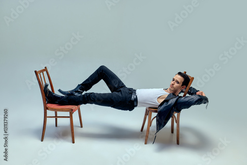 Foto Young man looks as rockstar, reclining between chairs in surreal fusion of movement and vintage aesthetic against muted studio backdrop