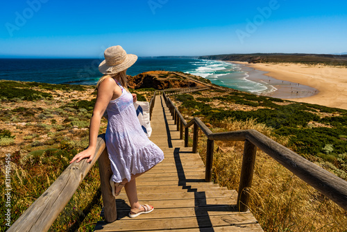 Mature woman tourist admiring picturesque view to cliff, ocean and sandy beach standing on wooden promenade in summertime. Side view. Bordeira beach panorama on Portugal coast