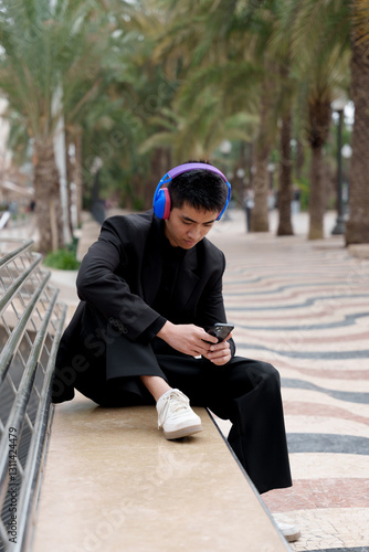 Young Asian man in stylish black suit listening to music with headphones while using his smartphone